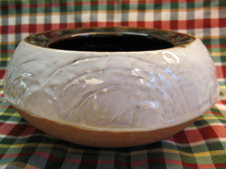 May include: A white ceramic bowl with a brown rim and a textured design. The bowl is sitting on a red and green plaid surface.