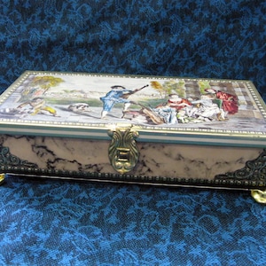 May include: A vintage tin box with a marbleized design and gold accents. The lid features a colorful illustration of people in a garden setting.