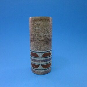 May include: A cylindrical ceramic vase with a textured, speckled brown surface. The vase features a decorative band of brown, blue, and purple geometric shapes near the base. The vase is approximately 8 inches tall and is set against a blue background.