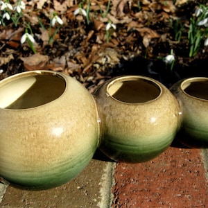 May include: Three round ceramic planters with a brown and green ombre glaze. The planters are arranged in a row on a brick surface.