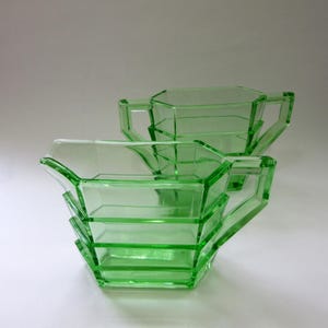May include: Two-piece set of green glass serving dishes. The set includes a sugar bowl and a creamer, both with geometric designs and handles. The glass is a vibrant green color, and the pieces are likely vintage.