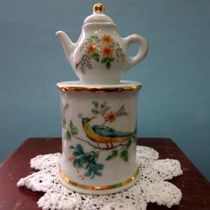 May include: A white ceramic teapot with orange floral accents sits atop a matching cylindrical container. The container features a painted bird design with yellow, blue, and green colors, and gold trim. The set is displayed on a white lace doily.