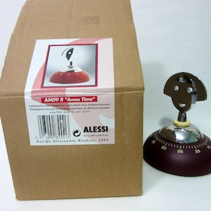 May include: A red and chrome-plated kitchen timer with a smiling face design. The timer has a circular base with numbers and a chrome head. The box next to it has the text "Anna Time".