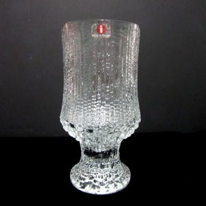 May include: Clear glass vase with a textured, bark-like pattern. The vase has a wide, cylindrical body and a pedestal base. A red and white sticker is visible near the top. The vase is on a black background.
