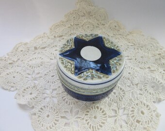 May include: A ceramic trinket box with a navy blue star design on the lid. The star has a white circle in the center. The box is round with blue and green stripes. The box is sitting on a white lace doily.