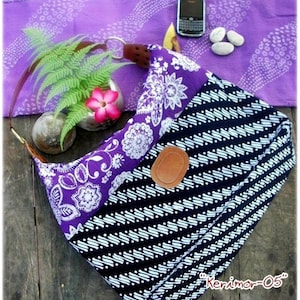 Op de afbeelding: Een paarse en zwarte stoffen tas met een wit bloemenpatroon en een bruine leren patch. De tas heeft een lange band en is gemaakt van een zacht, duurzaam materiaal. "Kerimar-05" is gedrukt op de bodem van de tas.