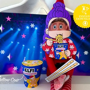 May include: A red and white Elf Superstar paper cutout with a purple wig and a face eating noodles. The cutout is holding a bowl of noodles and chopsticks. A blue and yellow cup with the text "Elfi Superstar" is also present. A background shows a stage with lights and stars.