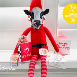 May include: A red elf wearing a cow mask holds a Chick-fil-A box with chicken nuggets. The elf is sitting on a white surface with a sign that says "Love Hugs and Chickie-Nuggs". There is another sign that says "I made a stop to eat mor chikin it was a pleasure!"