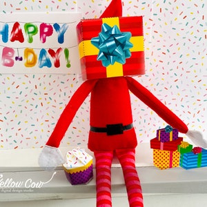 May include: A red elf doll wearing a red and yellow gift box as a hat. The elf is holding a purple cupcake and a stack of wrapped gifts. The background is a white surface with colourful confetti.