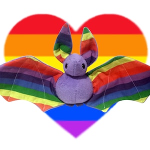 Peut inclure: Une chauve-souris en peluche violette avec des ailes arc-en-ciel, sur un fond de cœur arc-en-ciel.