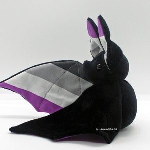 May include: A black plush bat with gray, white, and purple wings. The wings are patterned with the asexual pride flag.