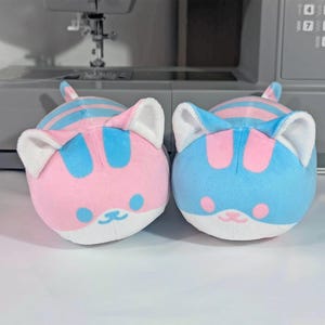May include: Two plush cat toys. One is pink with blue stripes, the other is blue with pink stripes. Both have white ears, a small tail, and a cute face with a pink nose and whiskers.