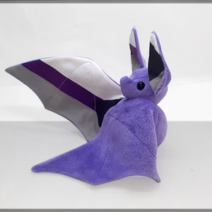 Könnte beinhalten: Lilafarbener Plüsch-Fledermaus mit grauen, weißen und lila Flügeln. Die Flügel sind mit den Farben der asexuellen Pride-Flagge gemustert.