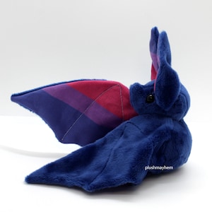 May include: A plush blue bat with a pink, purple and blue striped wing. The bat is lying on its side with its head turned to the right. The bat has black eyes and a small, pink nose. The text "plushmayhem" is visible on the bat's wing.