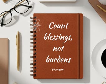 May include: A brown spiral-bound notebook with the text "Count blessings, not burdens" in white. The notebook is surrounded by a gold pen, black glasses, and other office supplies. The word "YTUMBLIN" is at the bottom.