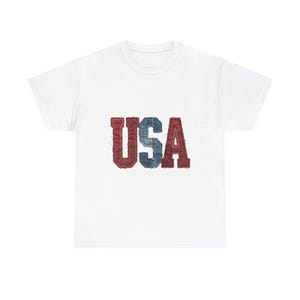 May include: A white short-sleeved t-shirt featuring the letters USA printed in the center. The letters have a textured, distressed appearance resembling red and blue denim fabric with visible stitching along the edges of each character.