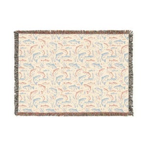 May include: Rectangular woven throw blanket with a cream background and a repeating pattern of salmon and trout in shades of blue and red. The blanket has a fringed edge.