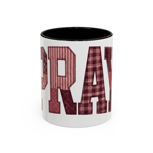 May include: White ceramic mug with the word "PRAY" in patchwork letters. The letters are various shades of red and burgundy, with a black interior. The mug is a standard size for coffee or tea.