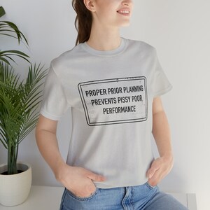 May include: A light gray t-shirt with a black and white sign that reads "PROPER PRIOR PLANNING PREVENTS PISSY POOR PERFORMANCE".