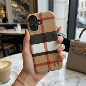 Könnte beinhalten: Beigefarbene Handyhülle mit rotem, schwarzem und weißem Karomuster. Die Hülle hat einen Kameraausschnitt und die Initialen "YB". Die Handyhülle wird von einer Person gehalten.