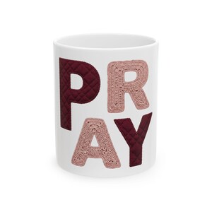 Può includere: Tazza in ceramica bianca con la scritta "PRAY" in bordeaux e rosa chiaro. Le lettere "P" e "Y" sono bordeaux, mentre "R" e "A" sono rosa chiaro. Le lettere hanno un aspetto trapuntato o all'uncinetto.