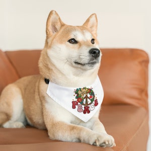 May include: A white dog wearing a white bandana with a red, green, and gold peace sign and the word "Peace" in a festive font. The bandana has a wreath of holly and berries around the peace sign.