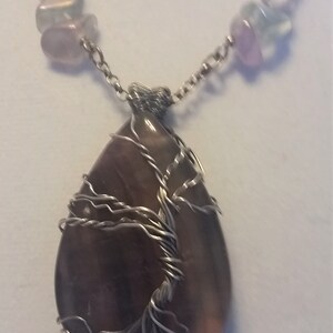 May include: A silver chain necklace with a teardrop-shaped pendant featuring a wire-wrapped tree design. The pendant is made of a dark gray stone with hints of purple and pink.
