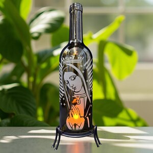 May include: A decorative wine bottle with a religious image etched on the front. The bottle is black and sits in a black metal stand. A small light inside the bottle illuminates the image. The background features green plants and a window.
