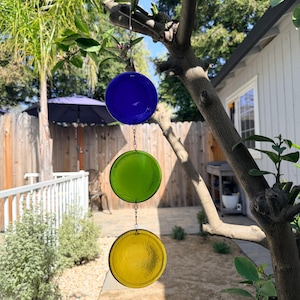 May include: A hanging outdoor decoration featuring three circular glass pieces in blue, green, and yellow. The glass discs are suspended by chains from a tree branch, creating a colorful accent for a garden or patio setting.