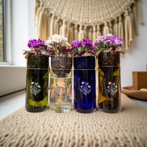 May include: Four glass planters with blooming African violets. The planters are made from repurposed bottles in green, clear, blue, and amber. Each bottle has a white floral design. The flowers have purple and white blooms and are set on a beige woven surface.
