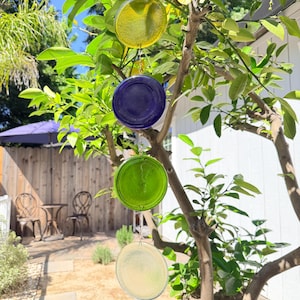 May include: A tree with green leaves and four hanging, circular glass ornaments in yellow, blue, green, and clear. The ornaments are suspended by chains. The background includes a wooden fence, a patio, and a blue umbrella.