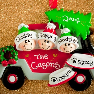 May include: A red and gray truck Christmas ornament featuring three smiling figures in hats labeled Daddy, Mommy, and Madilyn, with a green tree on top marked 2014. The truck side says The Casons, with two pet bowls labeled Roxy and Watson.