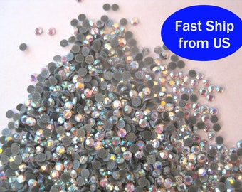 May include: A large pile of iridescent clear rhinestones, also known as crystals, with a silver backing. The rhinestones are scattered on a white surface.