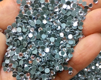 May include: A close-up of a hand holding a large quantity of small, clear, round rhinestones with silver backs. The rhinestones are scattered and overlapping.