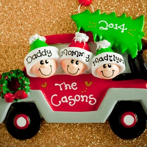 May include: A red and gray truck Christmas ornament featuring three smiling figures in hats labeled Daddy, Mommy, and Madilyn. A green Christmas tree on top displays the year 2014, and the side of the truck is inscribed with The Casons.