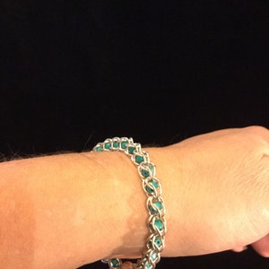May include: A silver-toned bracelet with turquoise-colored beads. The bracelet is made of interlocking metal links, creating a chain-like design. The turquoise beads are evenly spaced throughout the bracelet, adding a pop of color. The bracelet is worn on a wrist.