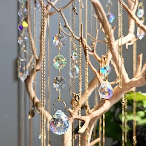 May include: A decorative jewelry display featuring a natural tree branch adorned with various necklaces and crystal pendants. The necklaces hang from the branches, showcasing clear, faceted crystals and gold-colored chains. The crystals reflect light, creating a sparkling effect.