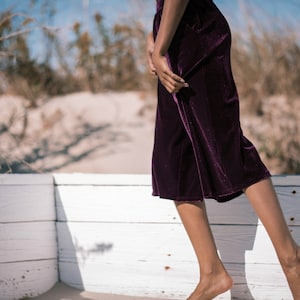 May include: A person wearing a pair of dark purple velvet culottes. The culottes are knee-length and have a soft, flowing texture. The background features a white wooden fence, sand, and dry plants.