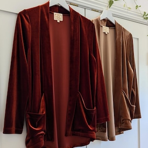 May include: Two velvet open-front jackets in shades of red-brown and tan hang on white hangers. Each jacket has two front pockets and a label at the neckline. The jackets are displayed against a white background.