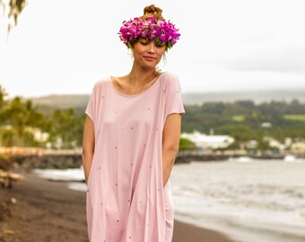 May include: A woman wearing a pink, short-sleeved dress with a floral crown on her head. She is standing on a sandy beach.