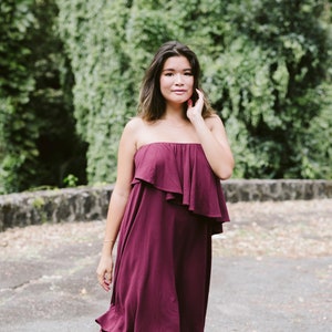 May include: A woman wearing a strapless burgundy dress with a ruffled neckline. The dress is knee-length and has a flowy silhouette.