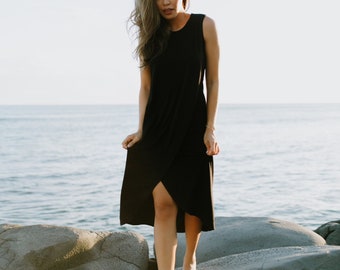 May include: A woman wearing a black sleeveless dress stands on a rocky beach. The dress has a wrap-style design and a slit in the front. The woman is looking at the camera.