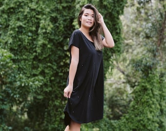 May include: A woman wearing a black, short-sleeved dress with a subtle sparkle. She is standing in front of a lush green background.