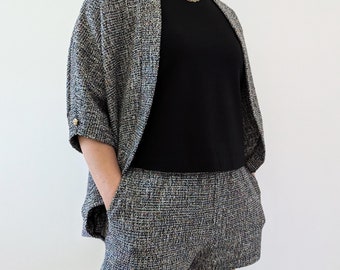 May include: A matching tweed set featuring a short-sleeved jacket and shorts. The jacket has gold buttons and the shorts have pockets. The set is made of a multi-colored tweed fabric, with black and white as the primary colors. A black top is worn underneath.