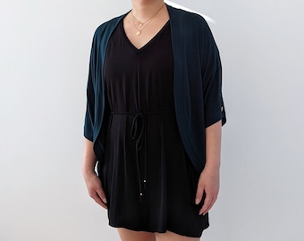May include: A black V-neck dress with a matching tie belt is worn under a dark teal, open-front, short-sleeved cardigan. The cardigan has a contrasting dark teal trim. A gold necklace is worn.