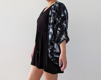 May include: A black short-sleeved romper with a drawstring waist paired with an open-front kimono featuring a black and white abstract forest or tree-like pattern. The ensemble consists of a solid black base layer and a patterned outer layer.