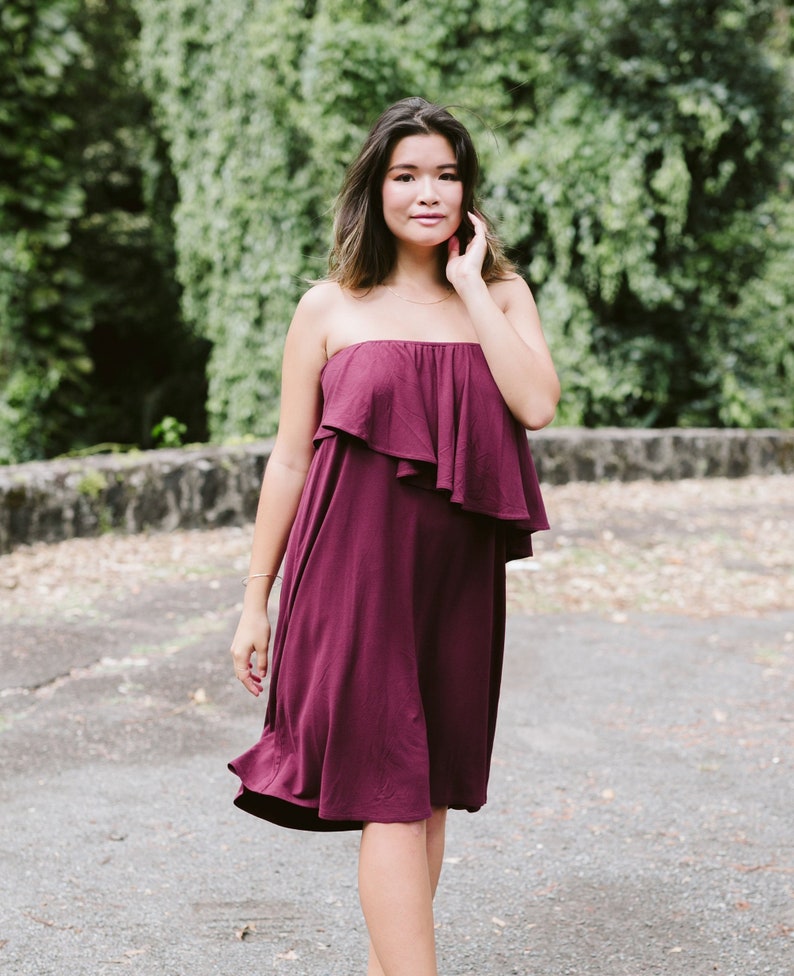 May include: A woman wearing a strapless burgundy dress with a ruffled neckline. The dress is knee-length and has a flowy silhouette.