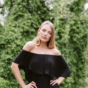 May include: A woman with long blonde hair is wearing a black off-the-shoulder dress with a ruffled neckline. She is standing in front of a green leafy background.
