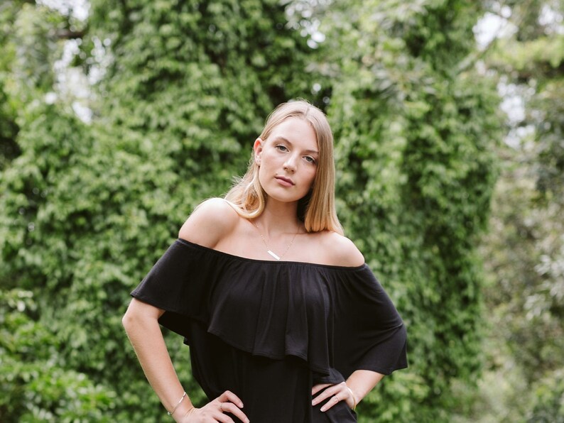 May include: A woman with long blonde hair is wearing a black off-the-shoulder dress with a ruffled neckline. She is standing in front of a green leafy background.