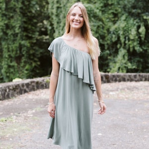 May include: A woman wearing a sage green one-shoulder dress with a ruffle detail. She is standing in front of a green leafy background.